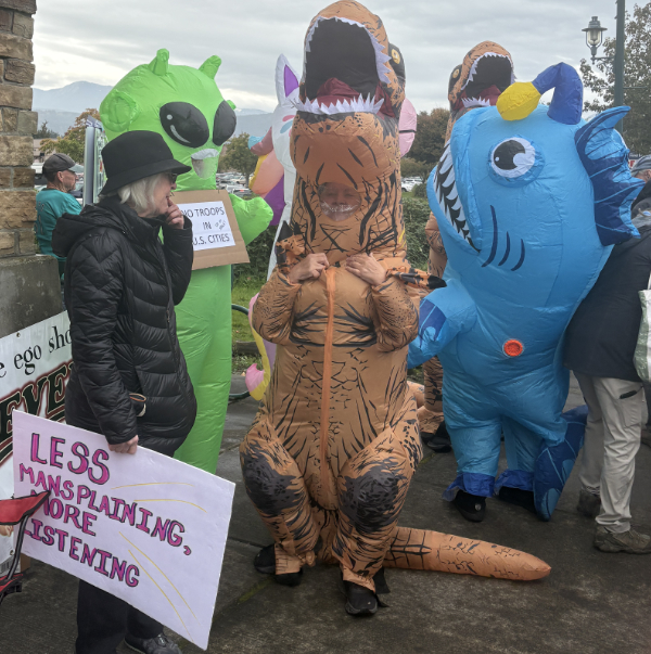 Get together with blow up dinosour, shark, and alien with human carrying sign Less Mansplaining More Listening